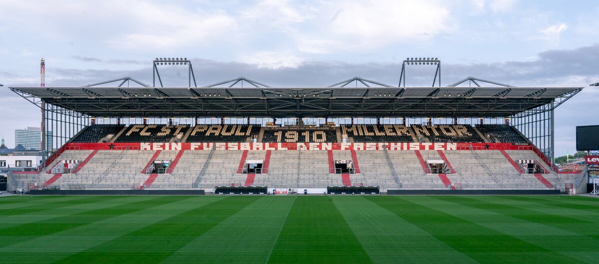 Foto einer überdachten Stadiontribüne. Davor ist ein akkurat gemähter Rasenfußballplatz. Auf der Tribüne steht: FC St. Pauli 1910 Millerntor. Kein Fußball den Faschisten.