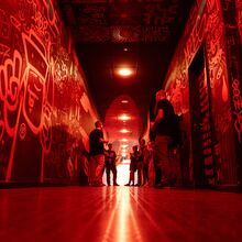 Foto eines langen Ganges, dessen Wände mit Graffiti besprüht sind. Das Foto ist vom Fußboden aus aufgenommen. Der Flur ist in rotes Licht getaucht. In der Mitte des Flures stehen Menschen.
