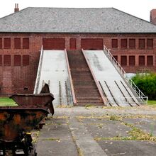 Rostige Eisenbahn-Loren vor einem Klinkergebäude. Zwei steile Rampen führen zu verschlossenen Toren im zweiten Stock des Gebäudes hinauf.