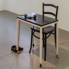 Einkarger Raum, in dem ein simpler, kleiner Holztisch sowie ein Stuhl steht. Auf dem Boden steht ein altes Wählscheibentelefon, dessen Hörer auf der Tischplatte liegt.