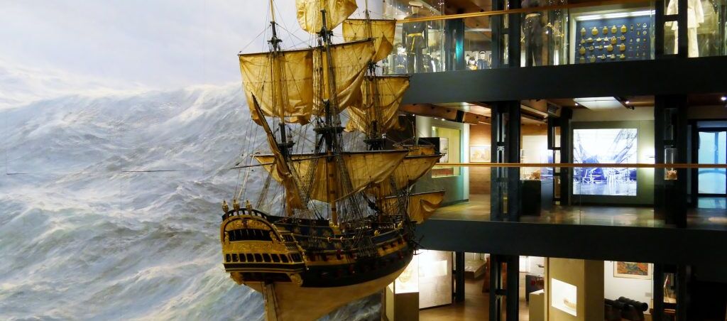 Modell eines historischen Dreimasters vor wogenden Wellen, die als Motivwand hinter dem Segelschiff zu sehen ist. Das Konstrukt befindet sich vor mehreren offene Etagen, die mit weiteren Ausstellungsstücken ausgestattet sind.