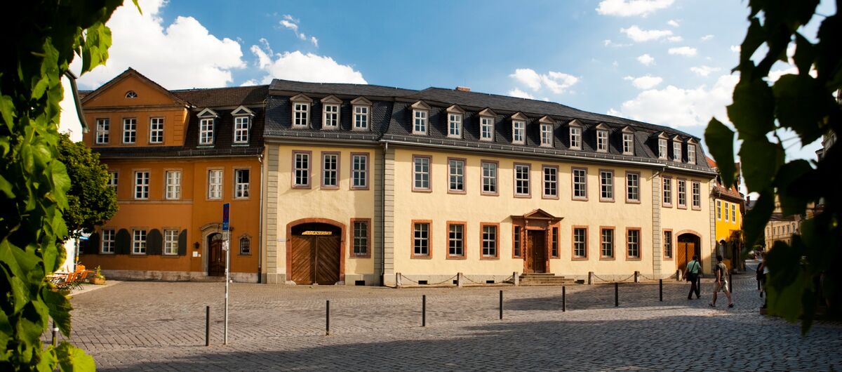 Foto einer dreistöckigen historischen Häuserzeile bestehend aus vier Häuserblöcken. Davor ist ein Platz aus Kopfsteinpflaster, in das Absperrfeiler eingelassen sind.
