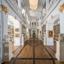 Foto vom historischen Bibliothekssaal mit breitem Holzgang unter kunstvoll verzierten Deckengewölbe. Die Wände zieren Regale mit goldenen Elementen, die Bücher, Porträtgemälden und Büsten enthalten.