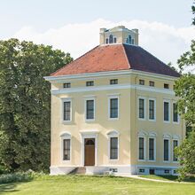 Foto des Schloss Luisium. Es ist ein historischer würfelartiger dreistöckiger Bau mit rotem Ziegeldach in einem Park. Auf dem Dach ist ein flaches Türmchen mit Fenstern draufgesetzt. Das Schloss ist hellgelb gestrichen, die Fenster weiß umrandet.
