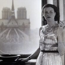 Schwarz-weiß Foto von Peggy Guggenheim. Sie steht im Kleid mit Perlenkette und Zigarette in ihrer linken gehobenen Hand vor einem Fenster in einem oberen Stockwerk. Ihre rchte Hand ruht auf einem Schrank vor dem Fenster.