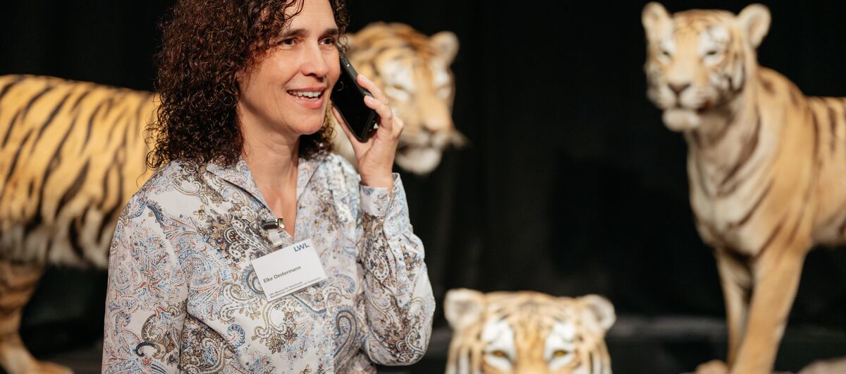 Foto einer Frau mittleren Alters, die ein Namensschild an der Bluse trägt und ein Handy an ihr linkes Ohr hält. Sie schaut freundlich. Im Hintergrund sind drei Tiger-Figuren vor einem schwarzen Hintergrund zu sehen.