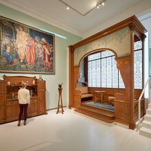 Foto eines Ausstellungsbereichs. Im Vordergrund an der Wand ein großer farbiger Wandteppich mit Szene der Geburt Jesu. Darunter eine Anrichte. Rechts daneben eine Holznische. Darin ein Sofa, runder Tisch und Stuhl. Die Möbel sind aus der Jugendstil-Zeit.