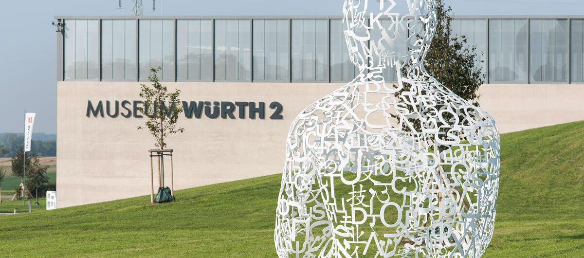 Foto der Außenansicht des Museum Würth 2. Vor dem modernen Gebäude befindet sich eine Wiese, die von rechts nach links abfällt. Auf der Wiese steht eine weiße Skulptur, die aus verschiedenen Schriftzeichen die obere Körperhälfte eines Menschen bildet.