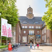 Menschen vor einem palastähnlichen Gebäude. Der rote Backsteinbau geht über 5 Etagen. Der Eingangsbereich ist über Treppen erreichbar, hat ein steinernes Tor und ist von Fenstern umrandet. Darüber ein Balkon. Auf dem Dach ist mittig ein Turm.