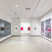 Ausstellungsansicht „Gerhard Richter. 100 Werke für Berlin“. In einem langen Raum hängen an der rechten weißen Wand drei große abstrakte Gemälde. Gegenüber auf der linken Seite hängen vier dunkle Spiegel. Geradeaus ist ein Durchgang zu einem weiteren Raum