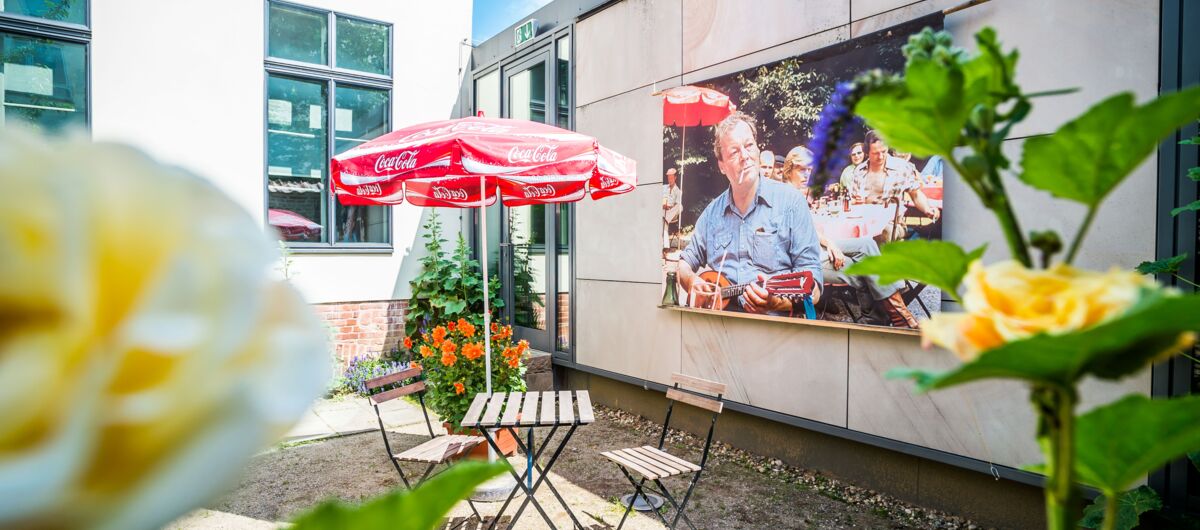 Innenhof in dem Rosen ranken, mittig steht ein Bistrotisch, Stühle und ein roter Sonnenschirm. Im Hintergrund ein großes Bild an der Wand: es zeigt Willy Brandt unter einem roten Sonnenschirm, mit Zigarette im Mund und Gitarre in der Hand sitzend.
