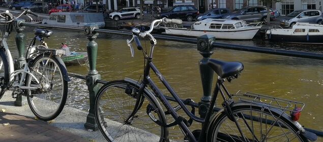Foto einer Gracht. Im Vordergrund stehen zwei Fahrräder angelehnt an einem Geländer. Auf der gegenüberliegenden Seite sind Boote festgemacht. Dahinter ist eine Hausreihe.