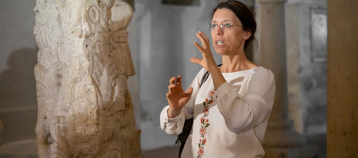 Foto in einer Kirche. Eine Frau spricht und gestikuliert vor ihrem Körper mit ihren Händen. Sie schaut links an der Kamera vorbei. Sie trägt ihr braunes Haar hochgesteckt und hat eine filigrane Brille auf.