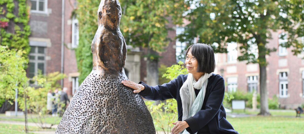 Foto einer Skulptur, die auf einem großen runden Sockel steht. auf dem Sockel sitzt eine Frau, die die Skulptur mit ihrer rechten Hand berührt. Diese Szene findet in einem Park statt. Im Hintergrund sind große Backsteinhäuser zu sehen.