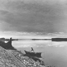 Schwarz-weiß Foto, auf dem ein einfaches kleines Holzboot am steinigen Rande eines stillen Sees liegt. Von dem Boot scheint ein Mensch herunterzuspringen. Der Blick über den See ist weit.