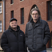 Foto von den Hinz & Kunzt Guides Chris und Uwe. Beide stehen nebeneinander vor einem Backsteinhaus, tragen warme Jacken und schauen mit einem freundlichen Blick in die Kamera..