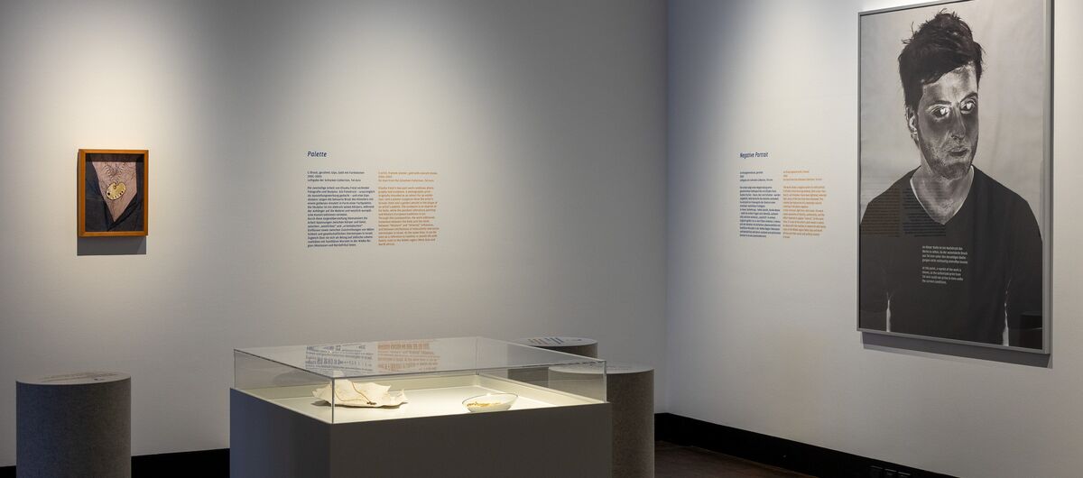 Foto eines Ausstellungsbereichs. An der rechten Wand hängt ein großes Portraitgemälde eines Mannes, an der linken Wand hängt ein kleines Gemälde - beide um Texte ergänzt. Im Raum steht eine Vitrine mit zwei Exponaten, die erleuchtet sind.