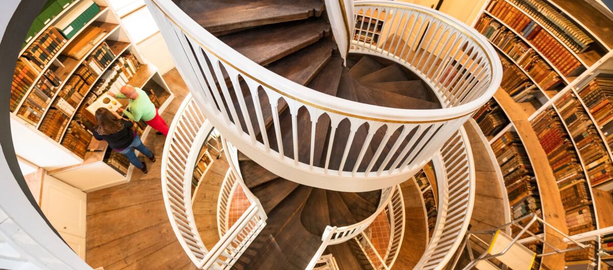 Foto mit Blick von oben nach unten in einem runden Turm. Zentral im Bild ist eine Wendeltreppe mit weißem Geländer und Treppenstufen aus dunklem Holz. An den gekrümmten Wänden stehen weiße Regale, in denen alte Bücher stehen.
