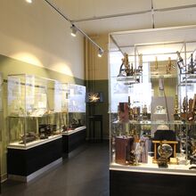 Foto eines Ausstellungsbereichs. In einem kleinen Raum mit Fenster an der Stirnseite stehen schmale Vitrinen an den Wänden und eine große Vitrine in der Mitte. In der mittigen sind historische Mikroskope zu sehen, in den anderen weitere technische Geräte.