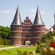 Foto des Holstentors bei Sonnenschein. Es ist ein Backsteinbau und besteht aus zwei großen Rundtürmen mit rundem spitzen Dach. Verbunden werden die Türme durch den Torbogen, über den sich weitere drei Geschosse ziehen. Davor ist eine Parkfläche mit Rasen.