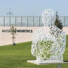 Foto der Außenansicht des Museum Würth 2. Vor dem modernen Gebäude befindet sich eine Wiese, die von rechts nach links abfällt. Auf der Wiese steht eine weiße Skulptur, die aus verschiedenen Schriftzeichen die obere Körperhälfte eines Menschen bildet.