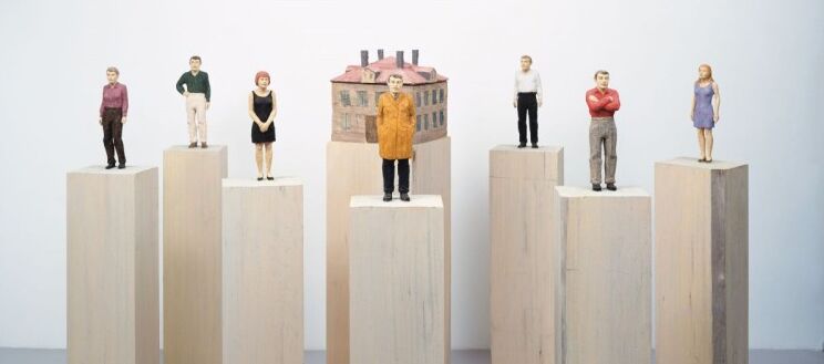 Foto von sieben Figuren und einem Haus aus bemaltem Holz, die jeweils auf einem Holzsockel stehen. Die Figuren stehen, haben aber unterschiedliche Körperhaltungen. Das dreigeschossige Haus ist würfelartig mit vier Schornsteinen.