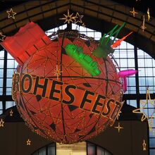 Foto von Weihnachtsbeleuchtung im Hamburger Hauptbahnhof: Eine rote Kugel mit dem Schriftzug Frohes Fest, die ca. zwei Meter im Durchmesser ist, hängt unter der Decke. Darauf sind weitere unterschiedliche Symbole Hamburgs.