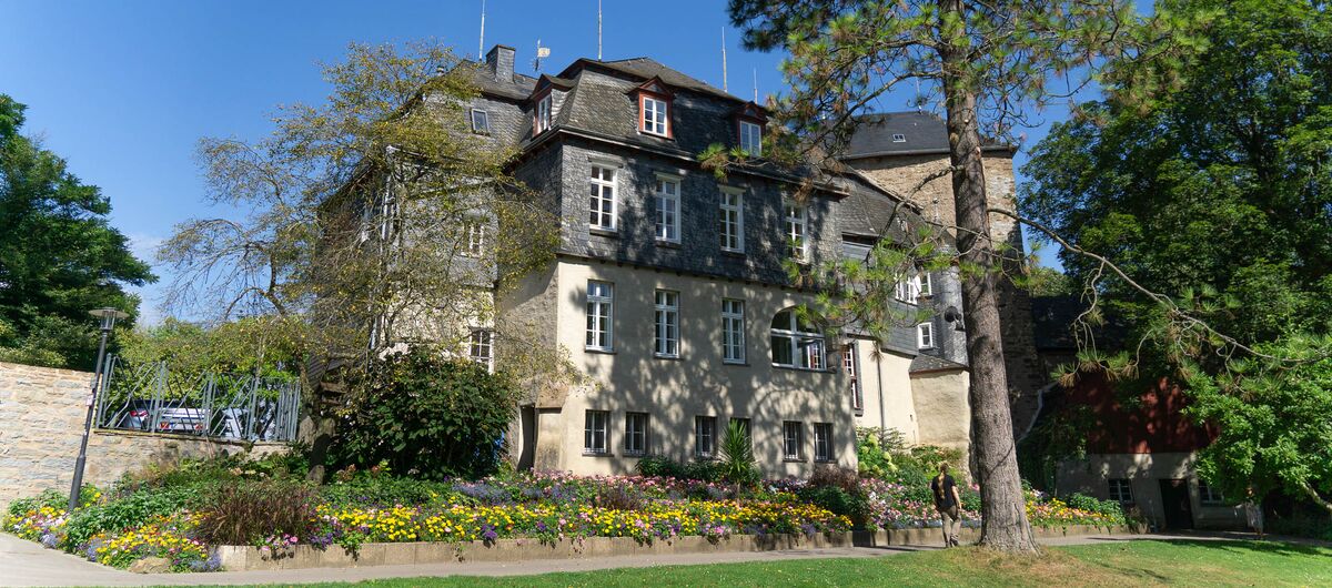 Foto eines viergeschossigen Schlosses, das verschachtelt mit Erkern und Türmchen gebaut wurde. Davor strahlt in bunten Farben ein üppiges Blumenbeet. Um das Haus stehen hohe Bäume. Im Vordergrund ist eine Rasenfläche. Der Himmel ist blau.