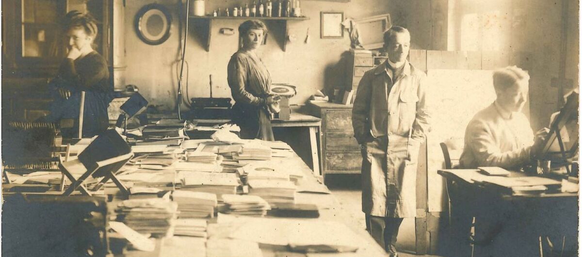 Schwarz-weiß Foto im Querformat eines Handwerksbetriebs. In dem Raum sind ein großer Tisch mit vielen Stapel Papier sowie weitere Arbeitsplätze und Schränke zu sehen. Zwei Frauen schauen direkt in die Kamera. Zwei Männer sind in ihr Tun vertieft.
