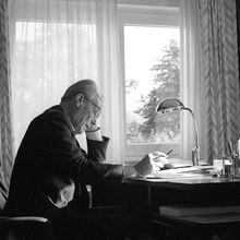 Schwarz-weiß Foto von Willy Brandt, der von der rechten Seite zu sehen an einem Schreibtisch sitzt. Er stützt seinen Kopf auf seinen linken auf den Schreibtisch aufgestützten Arm. Die rechte Hand liegt einen Stift haltend auf der Schreibtischplatte.