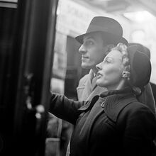 Schwarz-weiß Foto einer Frau und eines Mannes, die Ihren Blick auf etwas an der linken Fotoseite richten. Der rechte Arm der Frau ist leicht gehoben, alsob sie auf etwas zeigt. Beide sind mit Hut und Mantel fein gekleidet.