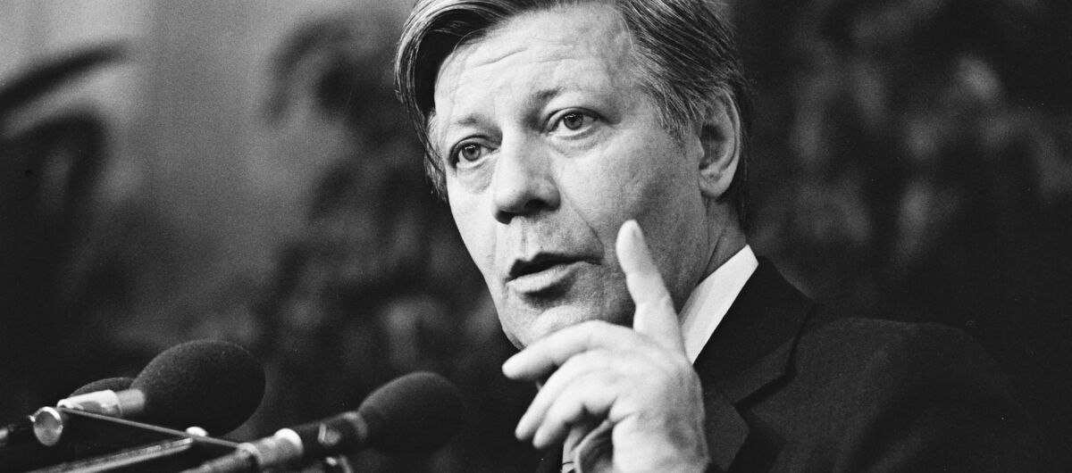 Helmut Schmidt steht mit erhobenen Zeigefinger an einem Rednerpult, von dem zwei Mikrofone in seine Richtung ausgerichtet sind.