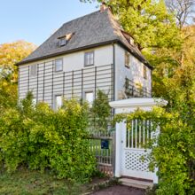 Weißes Haus in grünem Garten, zweigeschossig mit Schiefergedecktem Spitzdach. An den Wänden leere Rankhilfen, die Sprossenfenster sind mit Gardinen verhangen. Ein Zaun mit weißem Gartentor begrenzt das Grundstück, welches in warmen Sonnenlicht liegt.