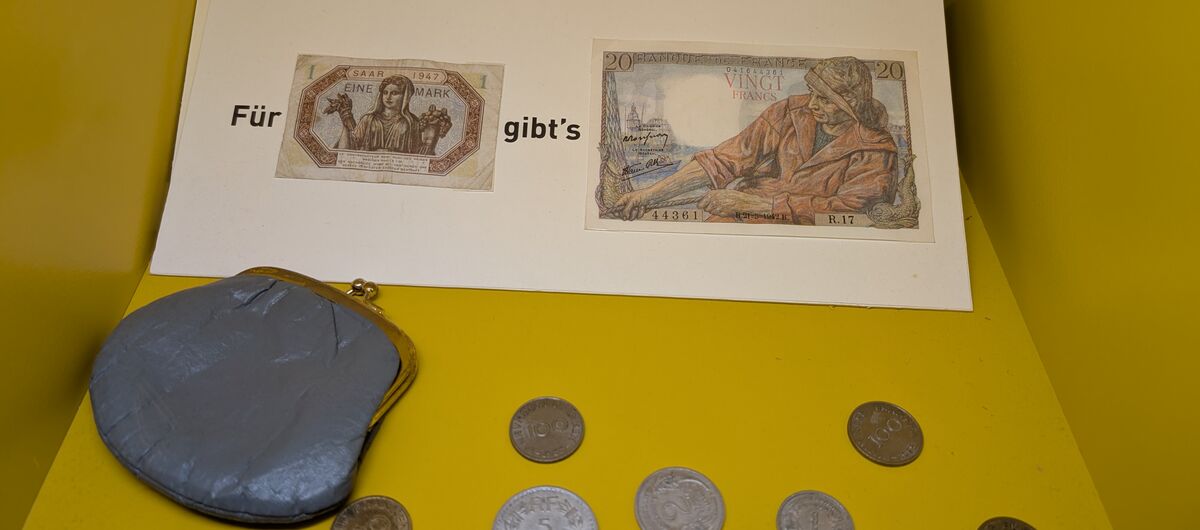 Foto von 18 Münzen unterschiedlicher Größe und Farbe. Links daneben liegt ein kleines graues Münzportemonnaie. Darüber liegt ein Papier auf dem ein kleiner und ein großer Geldschein kleben. Es steht: Für "kleiner Geldschein" gibt`s "großer Geldschein".