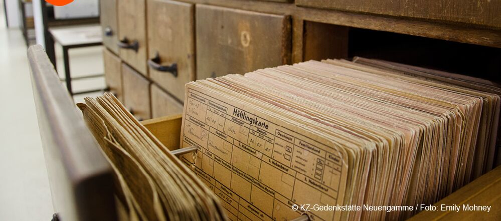 Foto eines seitlich fotografierten Holzschranks mit mehreren kleinen Schubladen. Die vorderste Schublade ist geöffnet. In Ihr stehen alte Häftlingskarten.