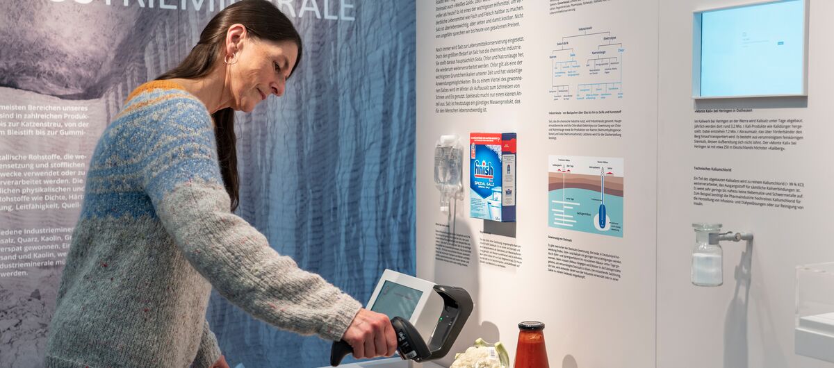 Foto eines Ausstellungsbereichs. Auf den Wänden sind Texte geschrieben, Abbildungen zu sehen und Exponate montiert. Auf einem Tisch davor stehen Lebensmittel unterschiedlicher Kategorien. Eine Frau hält einen Kassen-Scanner darüber.