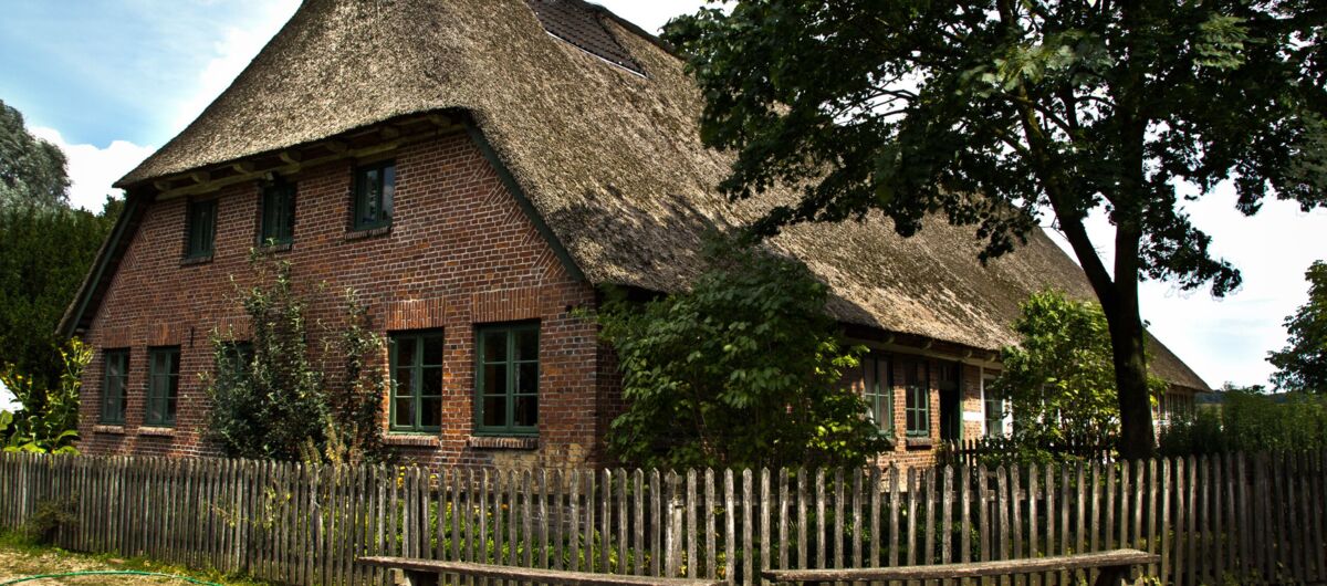 Foto eines geklinkerten Bauernhauses mit Reetdach und grünen Sprossenfenstern. Das Haus ist von einem Holzzaun eingefasst. Vor diesem stehen zwei einfache Holzbänke. Im Garten des Hauses stehen ein großer Baum und weitere Büsche.