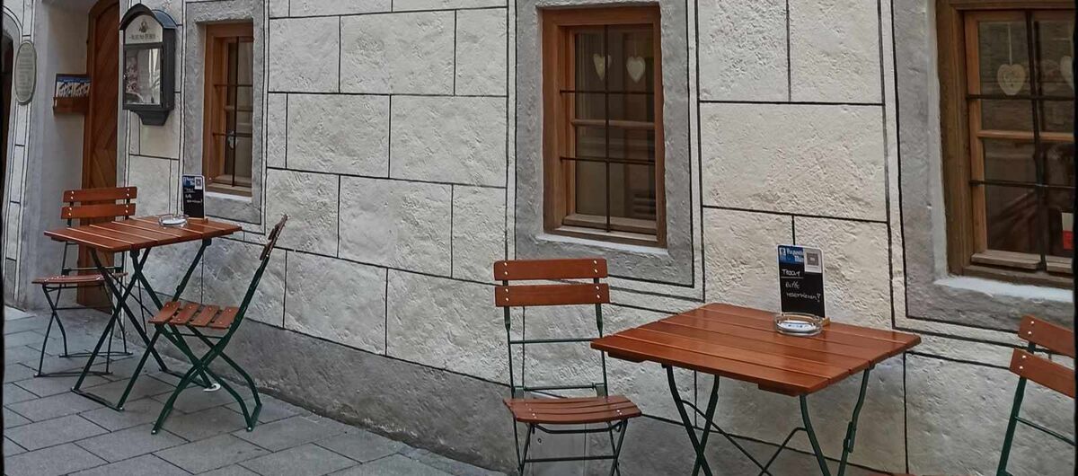 Foto eines Gebäudes. Davor stehen zwei kleine Klapptische mit je zwei Klappstühlen. An dem Haus ist ein Schild befestigt, auf dem "Bier Oktoberfest Museum" steht.