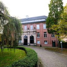 Historisches Backsteingebäude mit großen Fenstern und einem klassischen Eingangsbereich mit Säulen. Im Vordergrund befindet sich eine gepflegte Gartenanlage mit einem Kopfsteinpflasterweg, gesäumt von grünen Hecken und Bäumen.