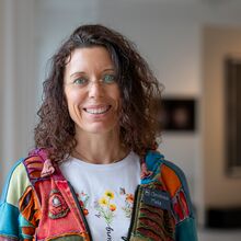 Foto in einem Ausstellungsraum. Eine freundlich lachende Frau ist bis zum Oberkörper erfasst. Sie trägt schulterlanges lockiges dunkles Haar, eine unscheinbare Brille, ein weißes T-Shirt mit Blumenmotiv und darüber eine bunte Patchwork-artig Kapuzenjacke.