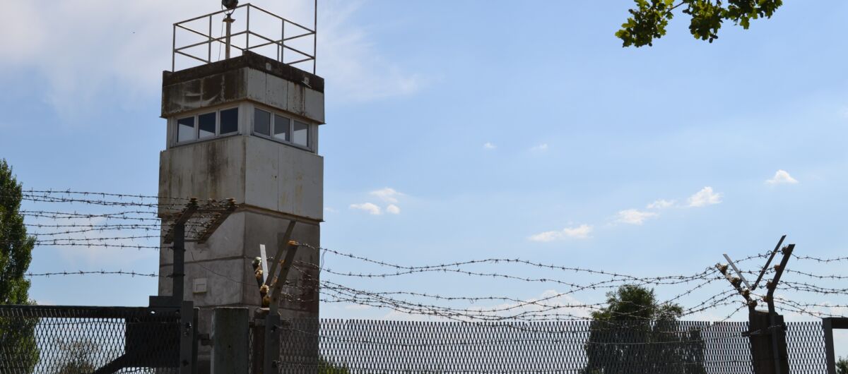Landschaft in dessen Vordergrund ein Grenzturm und hoher Zaun mit Stacheldraht ist.
