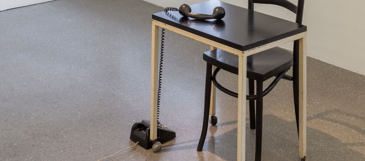 Einkarger Raum, in dem ein simpler, kleiner Holztisch sowie ein Stuhl steht. Auf dem Boden steht ein altes Wählscheibentelefon, dessen Hörer auf der Tischplatte liegt.