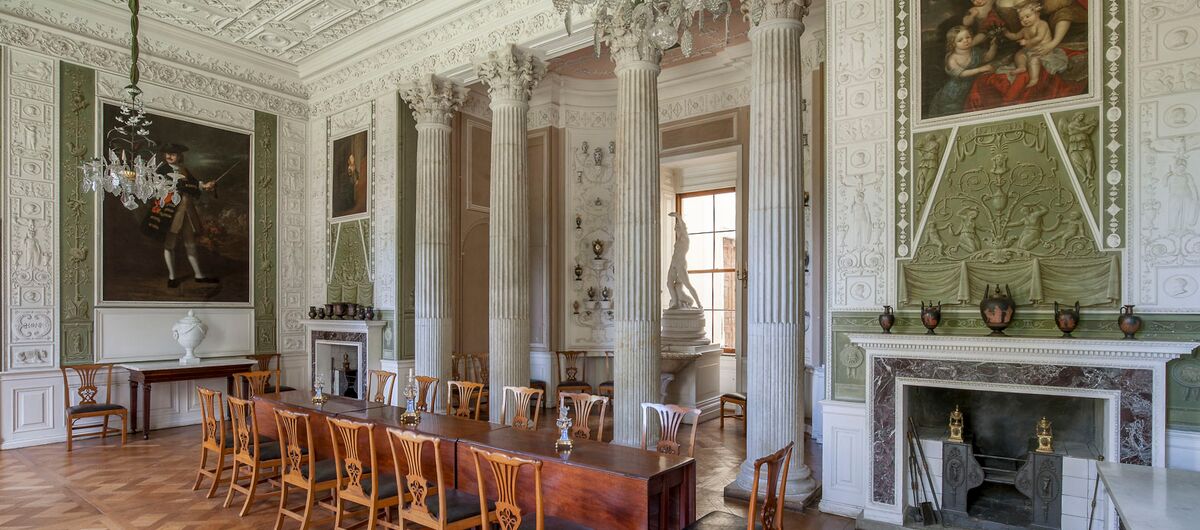 Foto des Speisesaals des Schloss Wörlitz. Kunstvoll verzierte weiße Wände werden durch einen von vier Säulen geschützten Erker sowie durch einen Kamin rechts daneben unterbrochen. Vor den Säulen sind 3 ausklappbare Holztische mit 14 Holzstühlen aufgebaut.