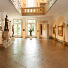 Foto der Diele des Behnhaus. Der Boden besteht aus alten großen speckigen Fliesen. Auf der linken Seite geht eine prachtvolle Treppe hoch, die am unteren Ende durch durch Statuen gesäumt ist. Auf der rechten Seite hängen drei große Gemälde an der Wand.