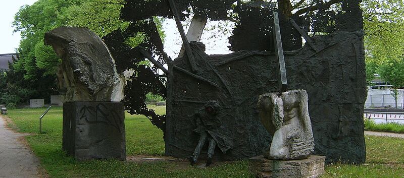 Foto eines Denkmals für den 2. Weltkrieg, das aus drei Teilen besteht. Zwei kleinere abstrakte Elemente aus Stein stehen je auf einem Steinsockel. Das größte Element ist schwarz, flächig und eine Person ragt mit Beinen und Mantel heraus, ebenso Stangen.