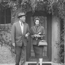 Schwarz-weiß Foto von Willy und Rut Brandt Ende der 50er Jahre. Sie stehen vor einer Haustür und schauen von der betrachtenden Person aus neutral nach rechts. Weinblätter und Efeu hängen über ihnen vom Haus hinunter.