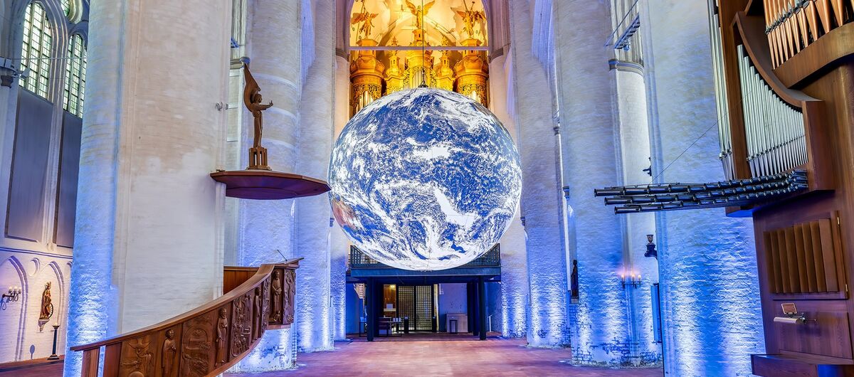 Foto des Innenraums der Hamburger Hauptkirche St. Katharinen. Zentral hängt die raumeinnehmende Installation "Gaia” - eine Erdkugel mit originalen NASA-Abbildungen und einem Durchmesser von sieben Metern. Die weißen Säulen werden blau angestrahlt.