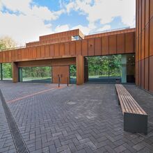 Außenansicht der Archäologischen Staatssammlung in München. Der moderne zweigeschossige Bau hat im Erdgeschoss eine dunkle Glasfassade. Die Eingangsflügeltür sowie der restliche Bau bestehen aus gerosteten Metallplatten.