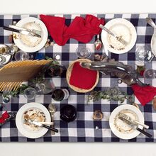 Foto von oben auf einen Esstisch nach dem Essen. Auf einem blau-weiß karierten Tischtuch stehen sechs schmutzige Teller mit Besteck, Servietten, Gläsern, Flaschen, Suppenteriene, Brotkorb, Gewürzen und wenig Deko aus Blättern.