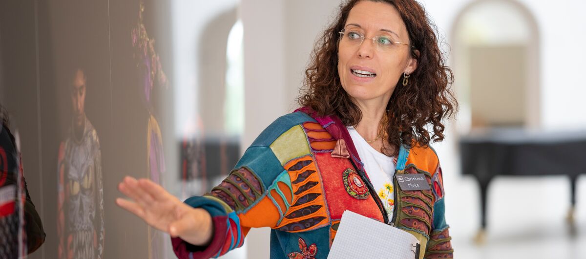 Foto eines Ausstellungsbereichs. Im Vordergrund ist eine Frau mit Zettel und Stift in ihrer linken Hand zu sehen, die spricht und mit ihrer rechten Hand auf ein großes Foto rechts neben ihr zeigt. Ihre Augen sind auf das Foto gerichtet.